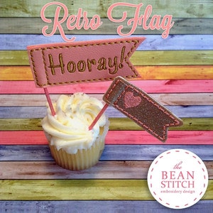 May include: A cupcake topped with white frosting and two pink and gold flags. The flags are decorated with the words "Hooray!" and a heart. The cupcake is sitting on a colorful wooden background. The text "Retro Flag" is written in pink and gold at the top of the image. The text "the BEAN STITCH embroidery design" is written in white on a pink circle at the bottom of the image.