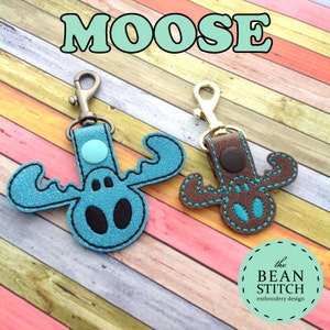 May include: Two moose-shaped keychains, one blue and one brown, with gold key rings. The blue moose has a turquoise button on its head. The text "MOOSE" is at the top of the image. The text "the BEAN STITCH embroidery design" is at the bottom of the image.