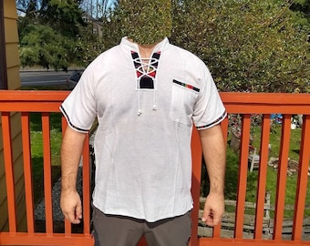 May include: A light-colored short-sleeved shirt with a decorative neckline and a small pocket. The shirt has black and red accents on the neckline and sleeves. The shirt is worn by a person standing on a wooden deck.