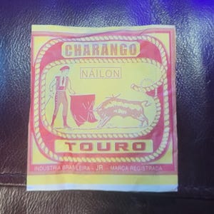 Könnte beinhalten: Eine gelb-rote Verpackung mit dem Wort "CHARANGO" oben. Die Verpackung zeigt eine Illustration einer Stierkampfszene mit einem Torero und einem Stier. Das Wort "NAILON" steht in der Mitte und "TOURO" unten.