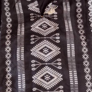 Może przedstawiać: Czarna tunika z białymi geometrycznymi wzorami. Tunika ma dekolt w kształcie litery V i szczeg&oacute;łowe białe wzory, w tym motywy diamentowe i liniowe. Tkanina wydaje się tkana, o teksturowanym wyglądzie.
