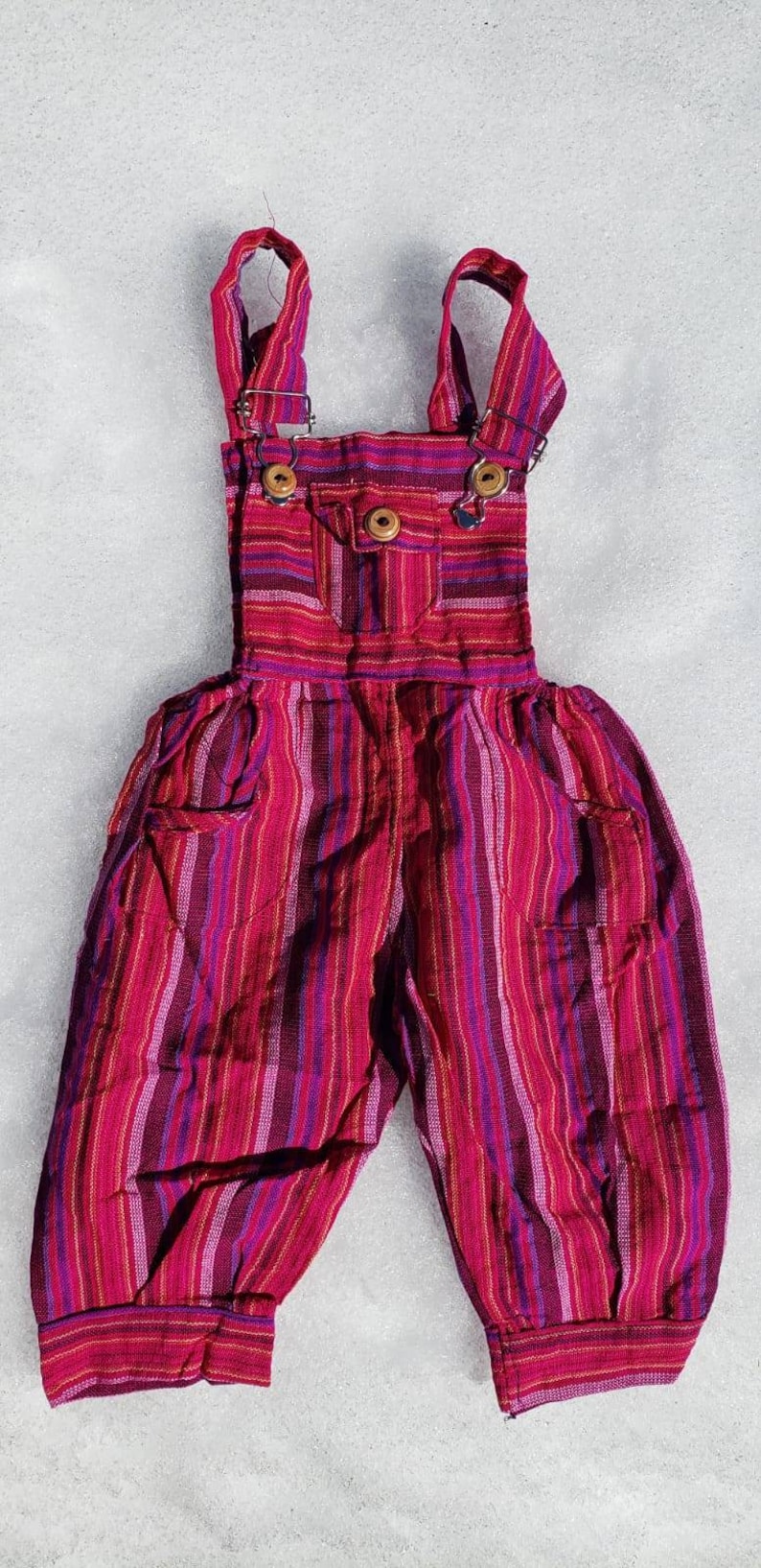 Puede incluir: Pantal&oacute;n vaquero rojo y morado a rayas con un peque&ntilde;o bolsillo en la parte delantera. El pantal&oacute;n vaquero tiene tirantes ajustables con botones de madera.