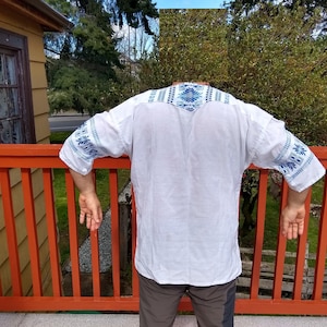 May include: White linen shirt with blue and white embroidered details on the sleeves and collar.