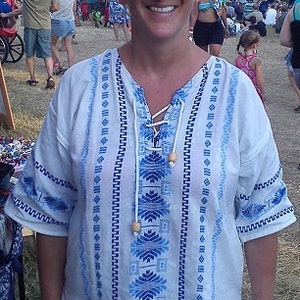 May include: A white embroidered blouse with blue and white geometric patterns. The blouse has a V-neck and a drawstring closure at the neckline. The blouse is made of a lightweight fabric and has a relaxed fit.