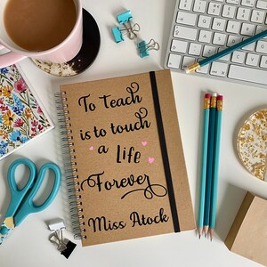 Op de afbeelding: Een bruin spiraalnotitieblok met een zwart lint-bladwijzer. De omslag heeft de tekst "To teach is to touch a life forever" in zwarte cursieve letters met twee roze hartjes. De naam "Miss Atock" staat in zwart onder de tekst.