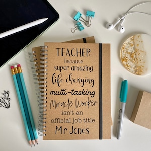Op de afbeelding: Een bruin notitieboek met een zwarte spiraalbinding. Het notitieboek heeft een zwart-wit tekstontwerp met de tekst "TEACHER because super amazing life changing multi-tasking Miracle Worker isn't an official job title Mr Jones".