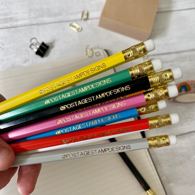 May include: A set of eight pencils with gold-colored accents and erasers. Each pencil is a different color and has the text "@postagestampdesigns" printed on it in gold lettering.