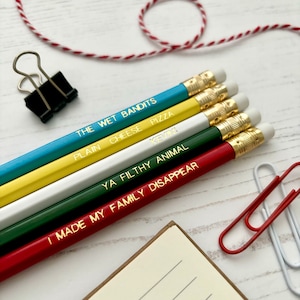 May include: Five wooden pencils with different colored bodies and gold-colored tips. Each pencil has a different funny phrase written on it in gold lettering. The phrases are "The Wet Bandits", "Plain Cheese Pizza", "Kemo", "Ya Filthy Animal", and "I Made My Family Disappear".