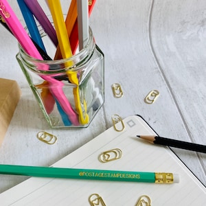 May include: A glass jar filled with colorful pencils with the text "POSTAGESTAMPDESIGNS" printed on each pencil. The jar is sitting on a white wooden surface. There are several gold paper clips with heart shapes on the surface.