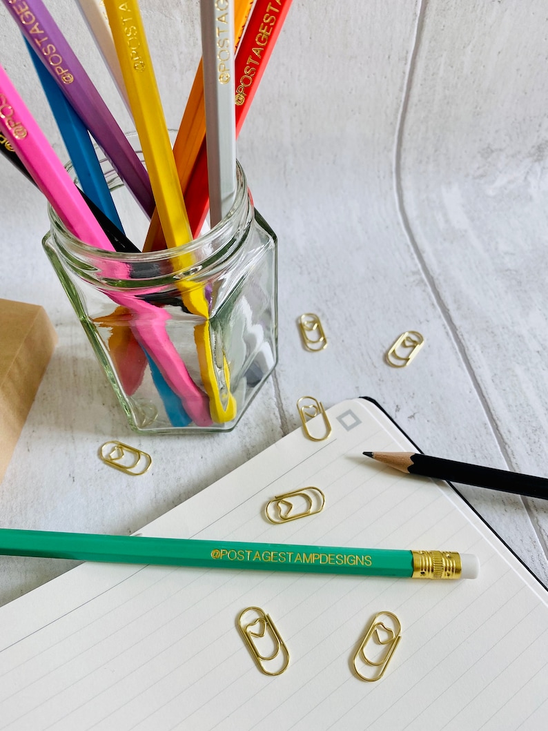 May include: A glass jar filled with colorful pencils with the text "POSTAGESTAMPDESIGNS" printed on each pencil. The jar is sitting on a white wooden surface. There are several gold paper clips with heart shapes on the surface.