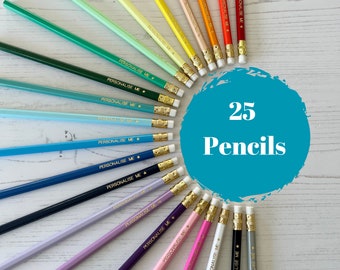 Può includere: Un set di 25 matite colorate disposte in cerchio su una superficie di legno bianco. Ogni matita ha una stella dorata e il testo "PERSONALISE ME" stampato su di essa.