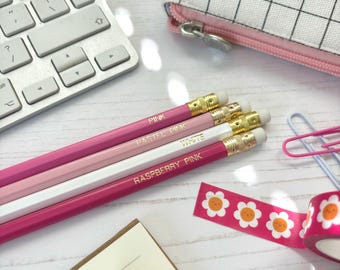Può includere: Primo piano di quattro matite rosa con estremità dorate, etichettate "Pink", "Pastel Pink", "White" e "Raspberry Pink". Sono su una superficie di legno bianco con una tastiera, un piccolo taccuino e nastro decorativo.