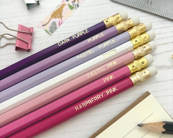Può includere: Una collezione di matite in varie tonalità di viola e rosa, ciascuna con un nome di colore dorato. Le matite hanno puntali in metallo dorato e gomme bianche. Sono visibili anche articoli di cancelleria come una graffetta, nastro adesivo e un righello.