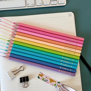 May include: A set of rainbow-coloured pencils, each labelled "ISABELLA" with a heart symbol, arranged on a lined notebook. A roll of patterned tape with cheetahs, and two binder clips are also visible. A white keyboard is in the background.