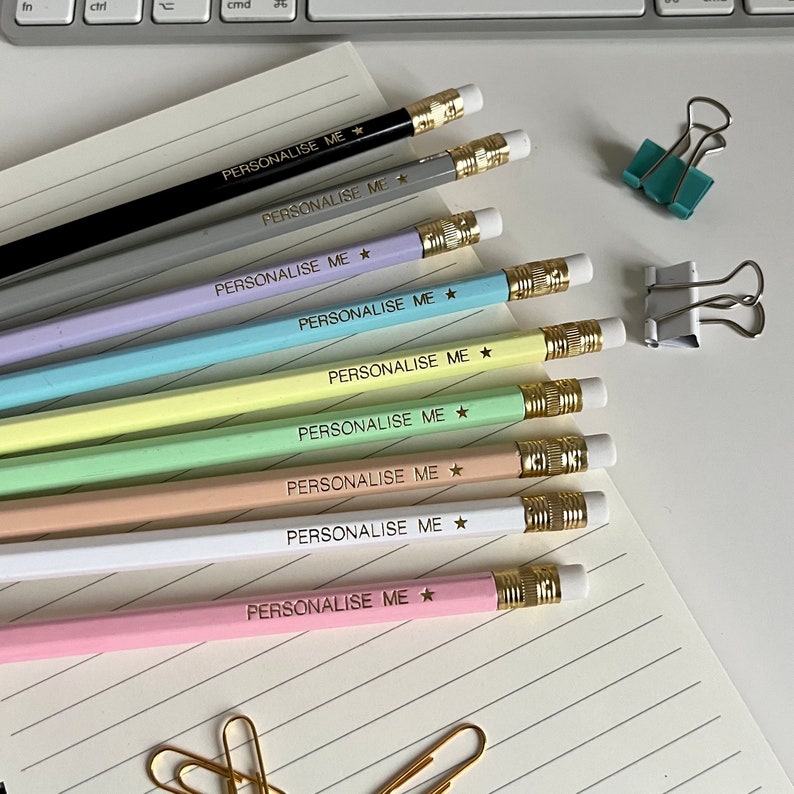 May include: A set of ten pencils with gold accents and a star design. The pencils are in various pastel colors, including pink, light blue, green, yellow, and gray. Each pencil has the words "PERSONALISE ME" printed on it.