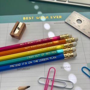 May include: A selection of rainbow-coloured pencils with gold text, such as "Not paid enough for this" and "Pretend it's on the lesson plan." A wooden sharpener and a metal ruler are on a planner with the words "Best Week Ever".