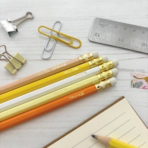 May include: A selection of pencils in shades of orange, yellow, and white, labelled "PASTEL ORANGE", "YELLOW", "WHITE", and "ORANGE", with erasers. Paper clips, a metal ruler, and a notepad are also present. The ruler is marked in inches.