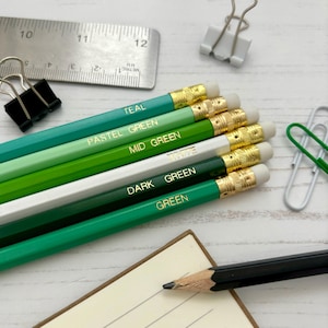 Op de afbeelding: Een verzameling potloden in verschillende tinten groen, turquoise en wit, elk met een gouden kleur naam. De potloden hebben goudkleurige metalen banden en witte gummen. Een metalen liniaal en paperclips zijn ook zichtbaar.