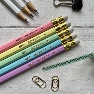 May include: Five pastel colored pencils with gold accents and erasers. Each pencil is labeled "MRS. BENNETT" with a gold star. The pencils are arranged in a rainbow order from pink to yellow.