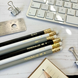 May include: A selection of pencils in black, white, and grey with gold lettering that reads "BLACK", "GREY", "WHITE", and "PERSONALIZE ME". The pencils have gold-coloured metal bands and white erasers. A keyboard, ruler, and paper clips are also visible.