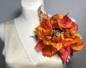 May include: A large, orange and pink fabric flower, possibly a brooch or corsage, is pinned to a white dress form. The flower has multiple layers of textured petals and a green center, with a few brown accents.