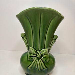 May include: A glossy forest green ceramic vase featuring a wide, flared fan-shaped top and a rounded base. The front of the vase is decorated with a molded relief of a tied ribbon bow, with two small, vertical decorative accents on each side.