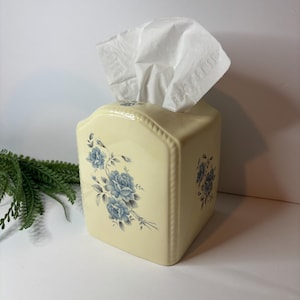 May include: A cream-colored ceramic tissue box cover with a rounded top edge and a beaded border. The front and side panels feature a delicate blue floral rose pattern. A white tissue is pulled through the top opening of the rectangular container.