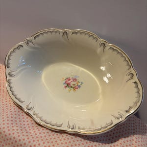 May include: An oval-shaped cream-coloured porcelain serving bowl with a scalloped edge. The rim features a decorative black line pattern with gold trim. A small, colourful floral bouquet of pink, yellow, and blue flowers is centred in the bowl.