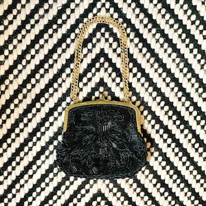 Puede incluir: Un bolso de noche negro de cuentas con una correa de cadena dorada. El bolso tiene un diseño floral y se cierra con un broche.