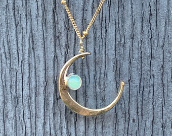 May include: A gold-colored necklace with a crescent moon pendant. The moon pendant has a light green stone in the center. The necklace chain is also gold and has small, round beads. The background is a weathered, gray wooden surface.
