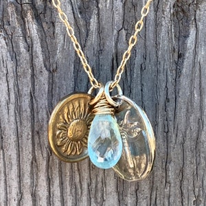 Puede incluir: Collar de cadena dorada con tres colgantes: un girasol, una libélula y una piedra preciosa azul claro en forma de lágrima.