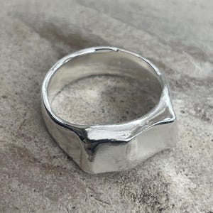 May include: A silver ring with a rectangular face and a smooth, polished finish. The ring has a thick band and a slightly textured appearance. The ring is shown on a neutral background.