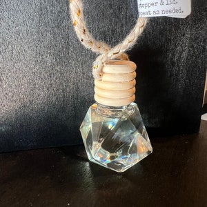 May include: A clear glass diamond-shaped car air freshener with a wooden stopper and a beige rope with gold accents. The air freshener has instructions on a white label that reads "DIFFUSER remove plastic topper, replace lid. tip upside down to wet top. replace plastic stopper & lid. repeat as needed."