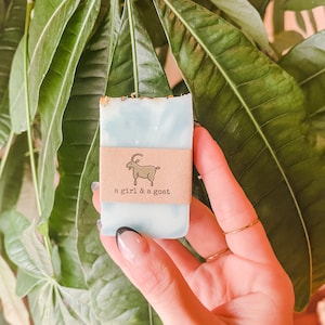 May include: A light blue soap bar with a white swirl design. The soap bar has a brown paper label with a white goat illustration and the text "a girl & a goat".