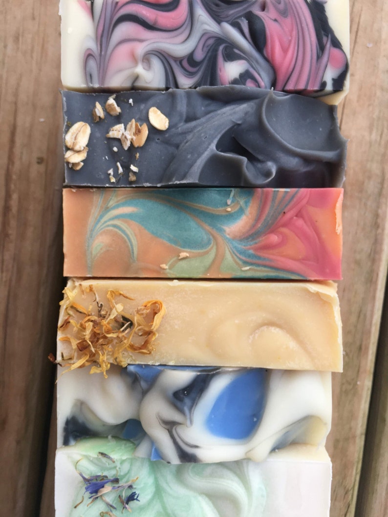 May include: A stack of six handmade soap bars with various colors and patterns. The top bar is pink and black with swirls, the second bar is gray with oatmeal flakes, the third bar is blue, green, and orange with swirls, the fourth bar is beige with dried flowers, the fifth bar is blue and white with swirls, and the bottom bar is green and white with swirls.