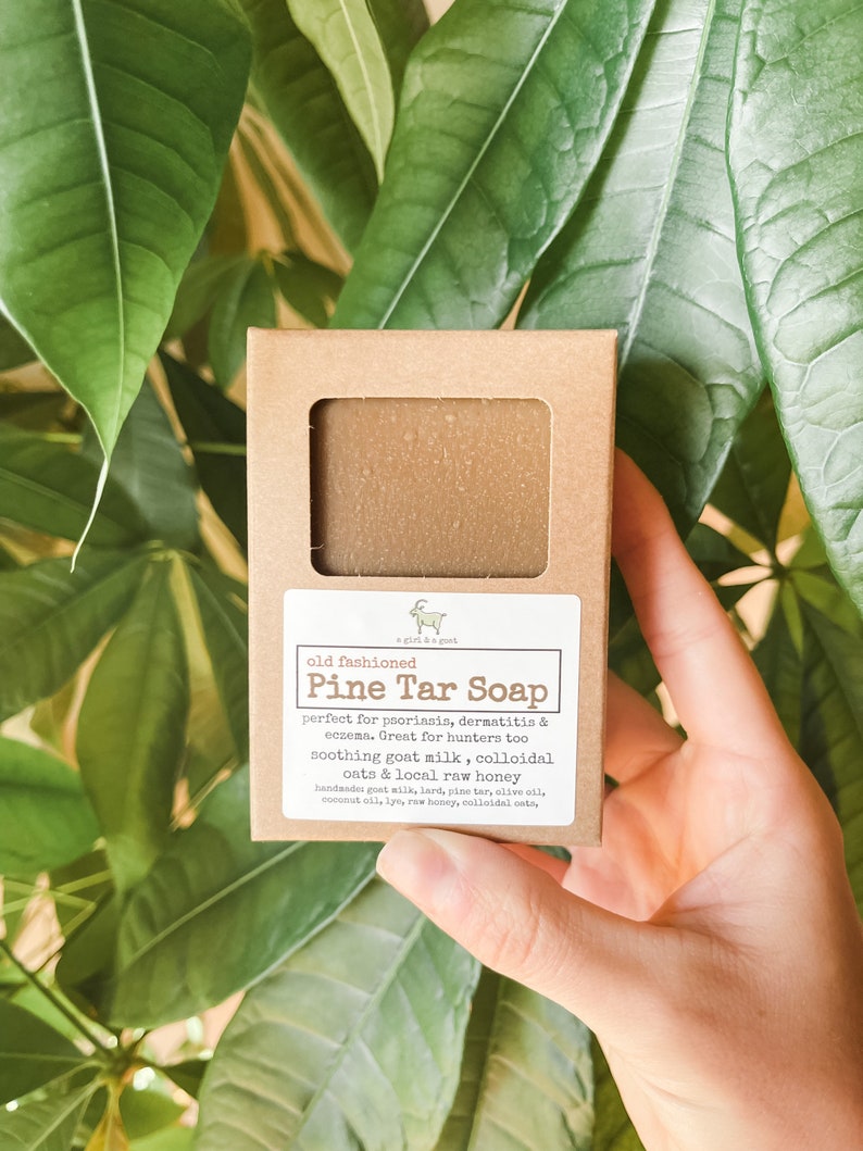 May include: A brown pine tar soap bar in a cardboard box with a window. The label says "Pine Tar Soap" and lists ingredients like goat milk and honey. The background features green leaves.