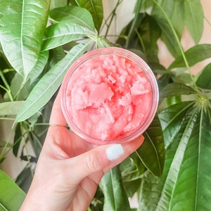 May include: A close-up of a pink sugar scrub in a clear jar. The scrub is thick and creamy, and it has a light pink color.