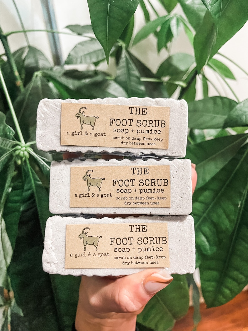 Może przedstawiać: Trzy białe kostki mydła z ilustracją kozy i tekstem "THE FOOT SCRUB soap + pumice scrub on damp feet. keep dry between uses" na każdej kostce.