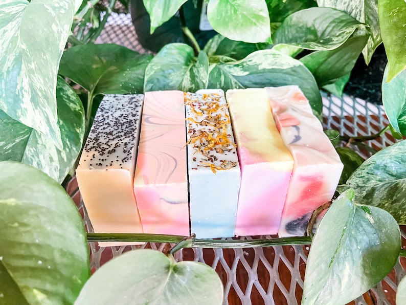 May include: Five bars of handmade soap, each with a different color and design. The bars are arranged in a row on a metal rack. The soap bars are a light beige with poppy seeds, a light pink with swirls, a light blue with dried flowers, a light pink with a yellow swirl, and a light pink with a dark blue swirl.