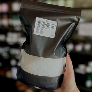 May include: A black resealable bag filled with white magnesium bath soak. The label reads "100% pure Magnesium Bath Soak" and lists the known benefits: reduce muscle tension, improved sleep, relaxed energy, detoxify skin, relieve pain, absorb minerals, silky soft water. Net wt. 32 oz.