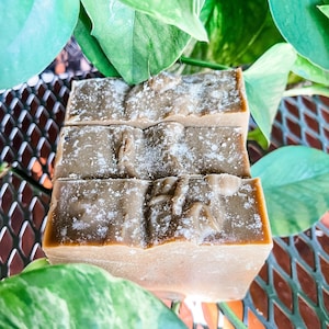 May include: Three bars of handmade soap, each with a light brown hue and a textured surface. The soap is dusted with a white powder and is surrounded by green leaves. The soap is displayed on a metal surface.