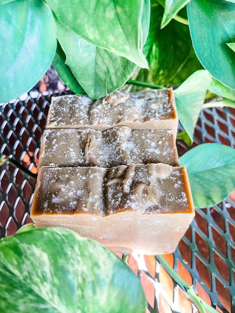 May include: Three bars of handmade soap, each with a light brown hue and a textured surface. The soap is dusted with a white powder and is surrounded by green leaves. The soap is displayed on a metal surface.