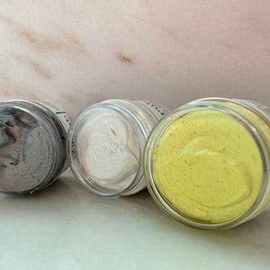 May include: Three small glass jars with lids containing different colored face masks. The jars are arranged in a row from left to right: gray, white, and yellow.