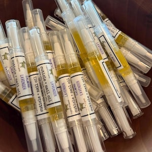 Op de afbeelding: Een close-up van een stapel doorzichtige plastic buisjes gevuld met een gele vloeistof. De buisjes hebben witte etiketten met de tekst "cuticle + nail oil" en "LAVENDER CALMING" erop gedrukt.