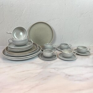 May include: A complete stoneware dinnerware set in a light gray color. The set includes dinner plates, salad plates, bowls, cups, and saucers. The plates and bowls have a simple, modern design, perfect for everyday use or special occasions.