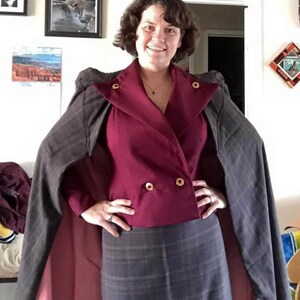 Op de afbeelding: Een vrouw draagt een bordeauxrode blazer met gouden knopen, een grijze geruite rok en een grijze en bordeauxrode cape.