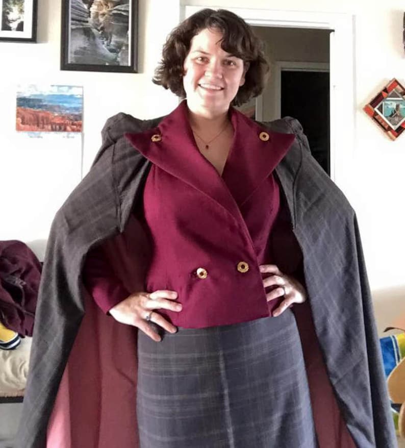 Op de afbeelding: Een vrouw draagt een bordeauxrode blazer met gouden knopen, een grijze geruite rok en een grijze en bordeauxrode cape.