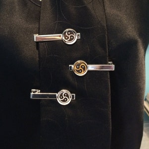 May include: Three silver-toned tie clips with a circular design featuring a black and gold symbol. The clips are attached to a black garment, creating a contrast between the shiny metal and the dark fabric. The design is centered on each clip.