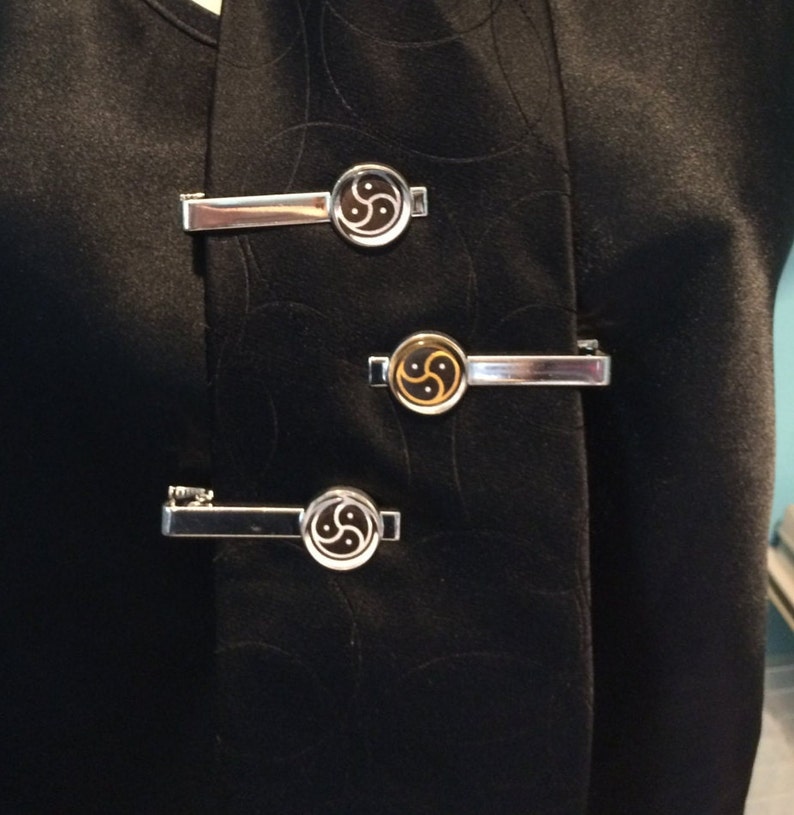 May include: Three silver-toned tie clips with a circular design featuring a black and gold symbol. The clips are attached to a black garment, creating a contrast between the shiny metal and the dark fabric. The design is centered on each clip.