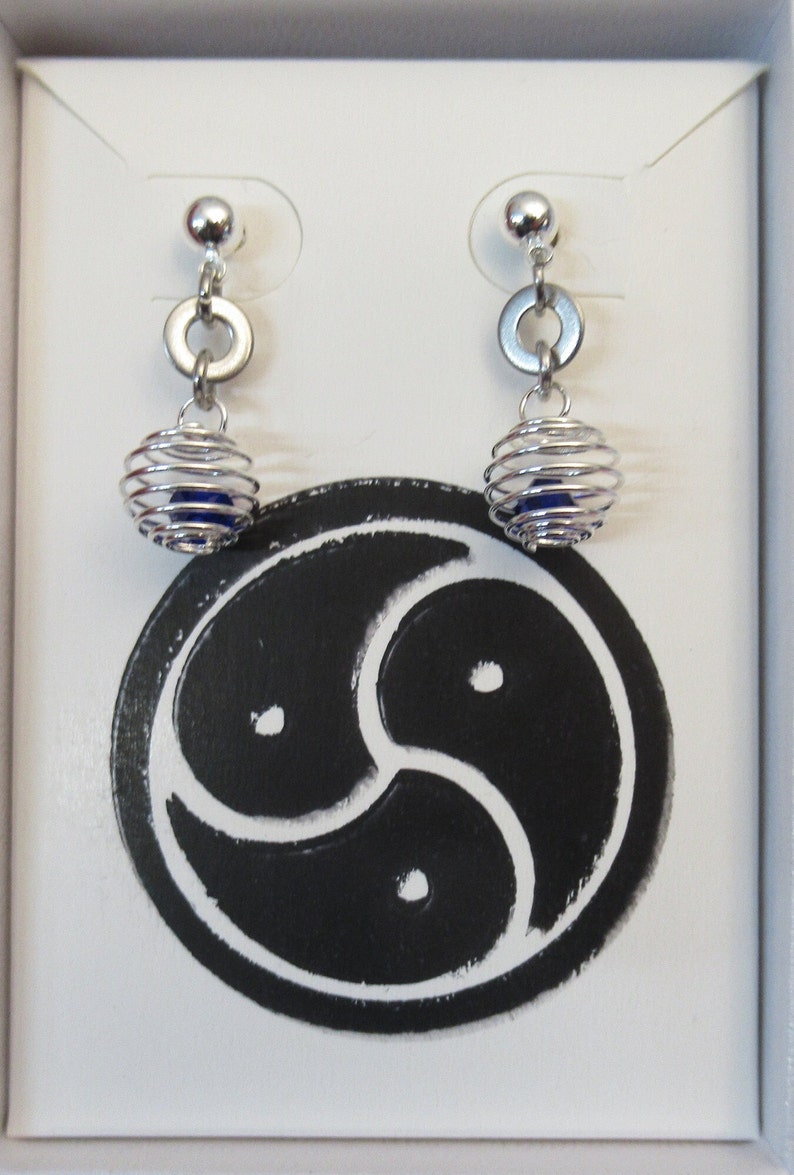 Puede incluir: Un par de pendientes de plata con una cuenta azul dentro de un dise&ntilde;o en forma de jaula. Los pendientes se muestran en una caja blanca con un s&iacute;mbolo negro.